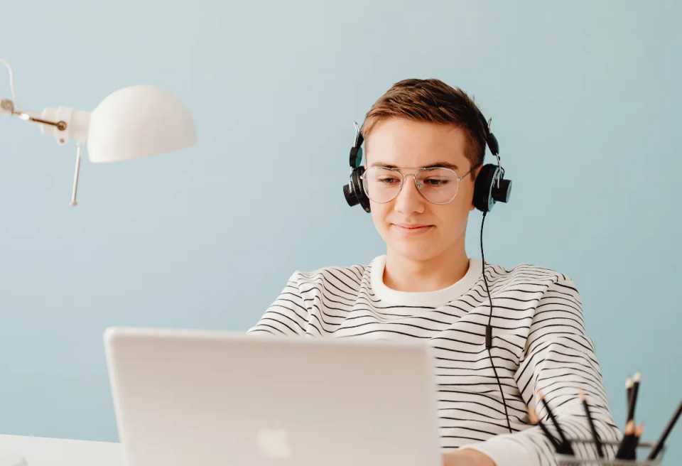 Young person wearing glasses and headphones, sitting at a desk with a laptop. They appear focused and content. The room has a soft blue wall and a white lamp.