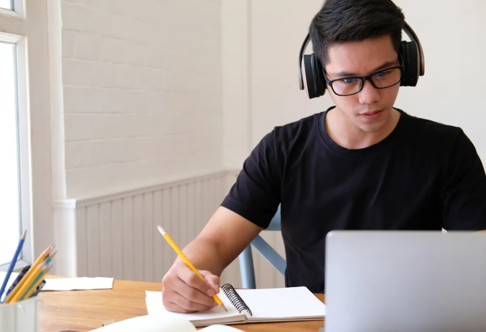 A focused young man wearing headphones and glasses sits at a table with a laptop, writing in a notebook. Daylight streams in from a nearby window.