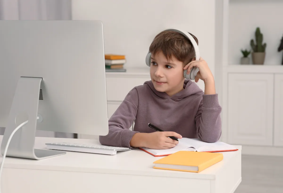 A child wearing headphones sits at a desk with a computer, taking notes in a notebook, surrounded by books and stationery.