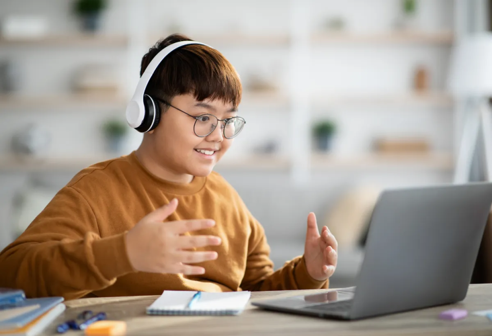 A person wearing headphones gestures while participating in a virtual meeting, with a laptop and notebook on a table in a bright room.