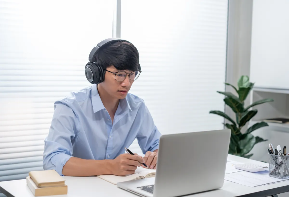 A person wearing headphones and glasses focuses intently on a laptop, pen in hand. They are seated at a desk with books and a plant in the background.