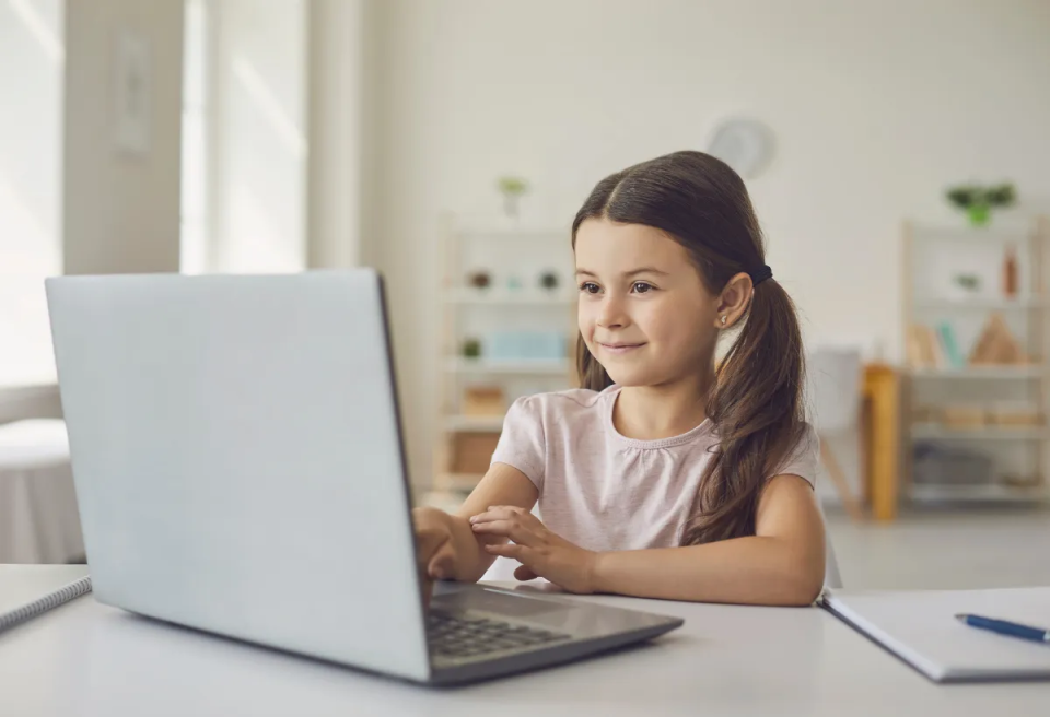 A girl in a light pink shirt sits at a desk, focusing on her laptop in a bright, modern room. A notepad and pen are nearby.