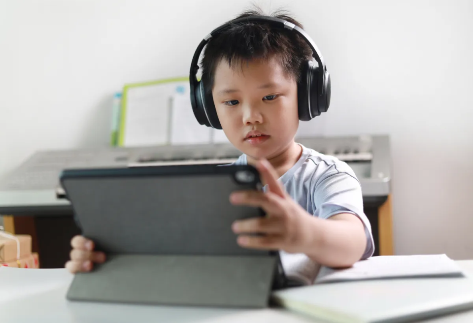 A child wearing headphones focuses on a tablet while seated at a desk with a keyboard in the background.