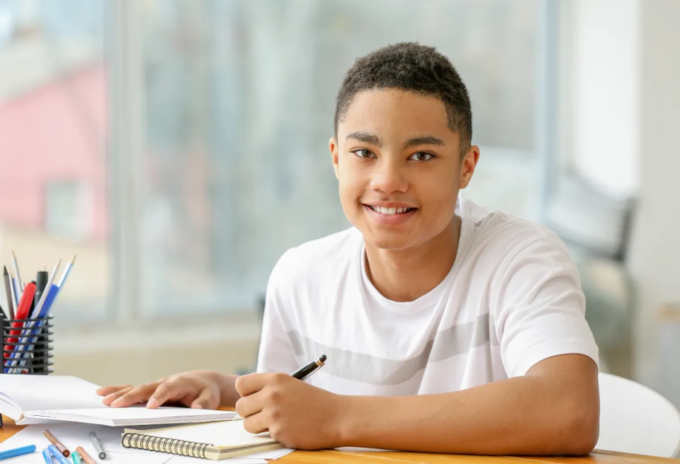 A student sits at a wooden desk with notebooks and pens, focused on writing while natural light streams through a window.